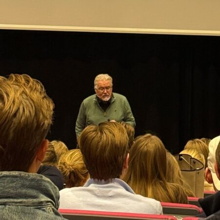 En foredragsholder står foran en gruppe tilhørere i et auditorium. På lerretet bak ham vises et bilde av Marie Curie med teksten «MARIE CURIE 1903». Publikum sitter i rekker og følger med på presentasjonen.