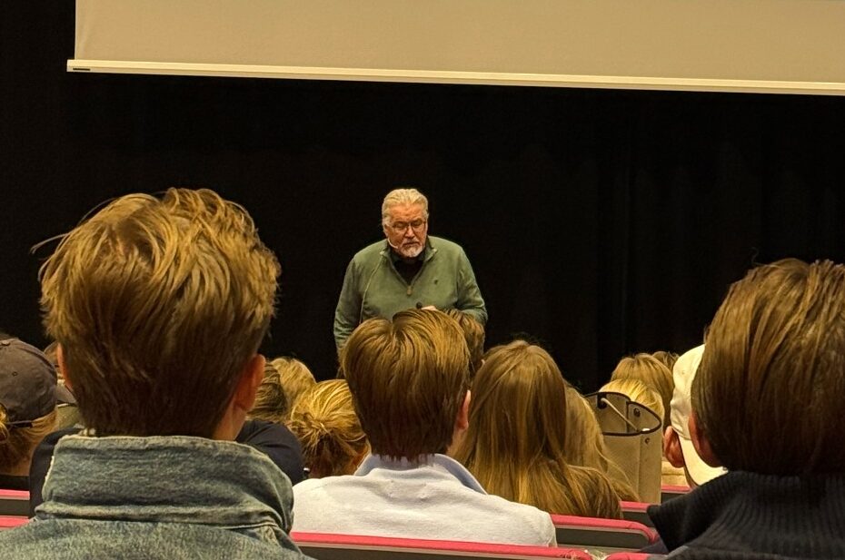 En foredragsholder står foran en gruppe tilhørere i et auditorium. På lerretet bak ham vises et bilde av Marie Curie med teksten «MARIE CURIE 1903». Publikum sitter i rekker og følger med på presentasjonen.